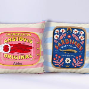 Könnte beinhalten: Zwei dekorative quadratische Kissen. Eines zeigt eine rosa Dose mit der Aufschrift "ANSJOVIS ORIGINAL" und "Abba". Das andere zeigt eine blaue Dose mit "SARDINES WILD CAUGHT IN OLIVE OIL". Beide haben gestreifte Hintergründe.