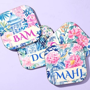 Puede incluir: Juego de cuatro posavasos cuadrados con esquinas redondeadas. Cada posavasos presenta un diseño floral con peonías rosas, pájaros azules y una pagoda. Los posavasos tienen texto en rosa y azul: BAM, DOG y MAHJ.