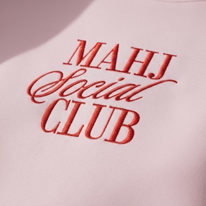 May include: Close-up of a light pink sweatshirt with the embroidered text "MAHJ Social CLUB" in red. The letters are in a mix of serif and script fonts. The fabric has a soft, textured appearance.