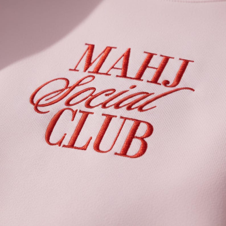 May include: Close-up of a light pink sweatshirt with the embroidered text "MAHJ Social CLUB" in red. The letters are in a mix of serif and script fonts. The fabric has a soft, textured appearance.