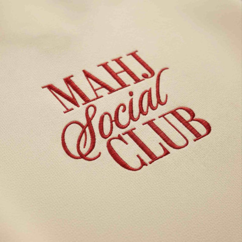 May include: Cream-colored fabric with embroidered text in red. The text reads "MAHJ Social CLUB" in a mix of serif and script fonts. The embroidery is detailed and the fabric has a subtle texture.