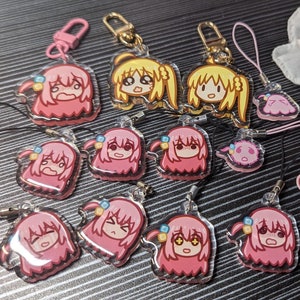 May include: A collection of acrylic keychains and charms featuring cartoon characters with pink or yellow hair, various facial expressions, and small blue hair accessories. The items include metal lobster clasps and black or pink cord loops.