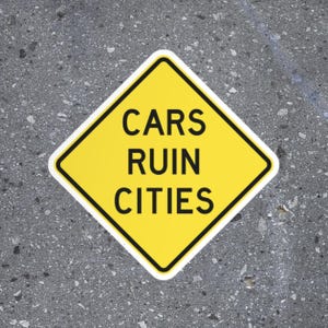 May include: A yellow diamond-shaped sign with a black border and the words "CARS RUIN CITIES" in bold black letters. The sign is set against a gray, textured background.