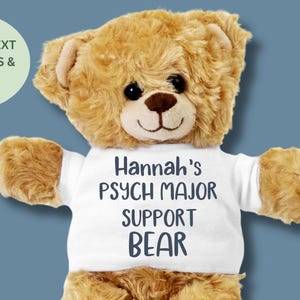 May include: A plush teddy bear with brown fur wearing a white t-shirt. The shirt reads "Hannah's PSYCH MAJOR SUPPORT BEAR" in dark blue lettering. The bear has a brown nose and black eyes.