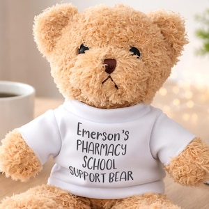 May include: A light brown plush teddy bear wearing a white t-shirt. The shirt features the black printed text Emerson's PHARMACY SCHOOL SUPPORT BEAR. The bear has dark eyes and a dark nose, with soft, textured fur covering its entire body.