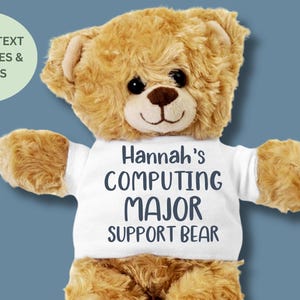 May include: A plush teddy bear with a white t-shirt that reads "Hannah's COMPUTING MAJOR SUPPORT BEAR." The bear is light brown with a dark brown nose and black eyes. A green circle with text is in the upper left corner.