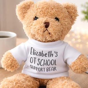 May include: A light brown plush teddy bear wearing a white t-shirt. The shirt features the dark gray printed text: Elizabeth's OT SCHOOL SUPPORT BEAR. The bear has dark eyes, a dark nose, and soft, textured fur, sitting in an upright position.