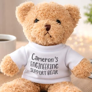 May include: A light brown plush teddy bear wearing a white t-shirt. The shirt features the black printed text Cameron's ENGINEERING SUPPORT BEAR. The bear has dark eyes, a dark brown nose, and soft, textured fur, sitting in an upright position.