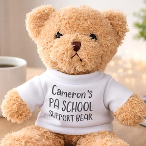 May include: A light brown plush teddy bear with dark eyes and a dark nose. The bear is wearing a white short-sleeved t-shirt with the black printed text Cameron's PA SCHOOL SUPPORT BEAR. The bear has soft, textured fur and sits in an upright position.
