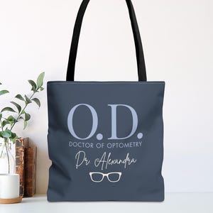 May include: Navy blue tote bag with black handles. The bag features the text "O.D. DOCTOR OF OPTOMETRY" in light blue, along with the name "Dr. Alexandra" and a pair of eyeglasses.
