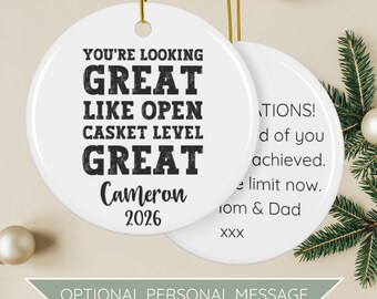 May include: A round white ceramic ornament hanging by a gold string. The front features black text reading: YOU'RE LOOKING GREAT LIKE OPEN CASKET LEVEL GREAT, Cameron, 2026. The back shows a personal message from Mom & Dad with the text XXX.