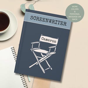 May include: A dark blue notebook with a light blue horizontal band at the top featuring the word SCREENWRITER in black text. Below, a white line drawing of a director's chair displays the name Cameron on the backrest in black lettering.
