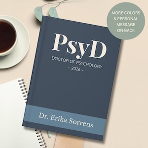 May include: A navy blue hardcover journal with the text "PsyD DOCTOR OF PSYCHOLOGY - 2026-" in white. The bottom features a light blue band with "Dr. Erika Sorrens" printed on it. A green circle with text is in the upper right corner. A cup of coffee and a notebook are in the background.
