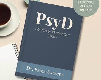 May include: A navy blue hardcover journal with the text "PsyD DOCTOR OF PSYCHOLOGY - 2026-" in white. The bottom features a light blue band with "Dr. Erika Sorrens" printed on it. A green circle with text is in the upper right corner. A cup of coffee and a notebook are in the background.