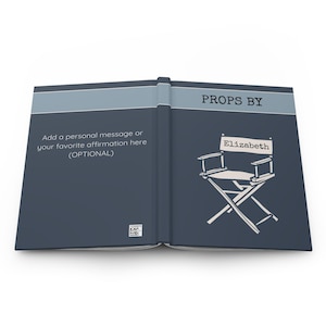 May include: Open journal with a dark blue cover and a light blue stripe. The right side features a director's chair illustration with the name "Elizabeth." Text on the left reads "Add a personal message or your favorite affirmation here (OPTIONAL)." The top reads "PROPS BY."