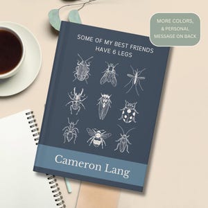 May include: A dark blue hardcover journal featuring white line art illustrations of nine different insects. The text reads SOME OF MY BEST FRIENDS HAVE 6 LEGS and the name Cameron Lang. A small box in the corner states MORE COLORS, & PERSONAL MESSAGE ON BACK.