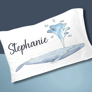 May include: White pillowcase featuring a watercolor illustration of a blue whale spouting water. The name "Stephanie" is written in a cursive font above the whale. The design is on a white background.