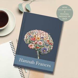 May include: A navy blue journal with a floral brain design. The brain is filled with colorful flowers. The journal has a light blue band with the name "Hannah Frances" printed on it. A coffee cup and notepad are in the background.