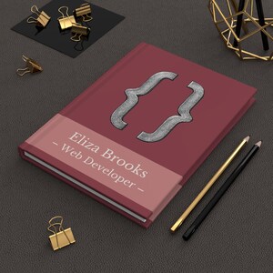 May include: A maroon notebook featuring a silver bracket design and the text "Eliza Brooks - Web Developer -". Gold and black pencils and gold binder clips are arranged around the notebook on a dark grey surface.