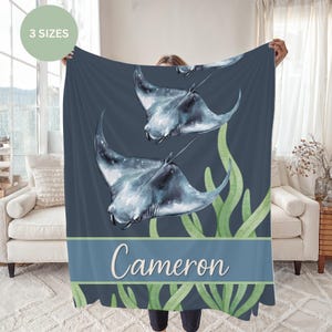 May include: A dark blue fleece blanket featuring a watercolor illustration of two manta rays swimming above green seaweed. A horizontal light blue band at the bottom contains the name Cameron in white script. A circular badge says 3 SIZES.