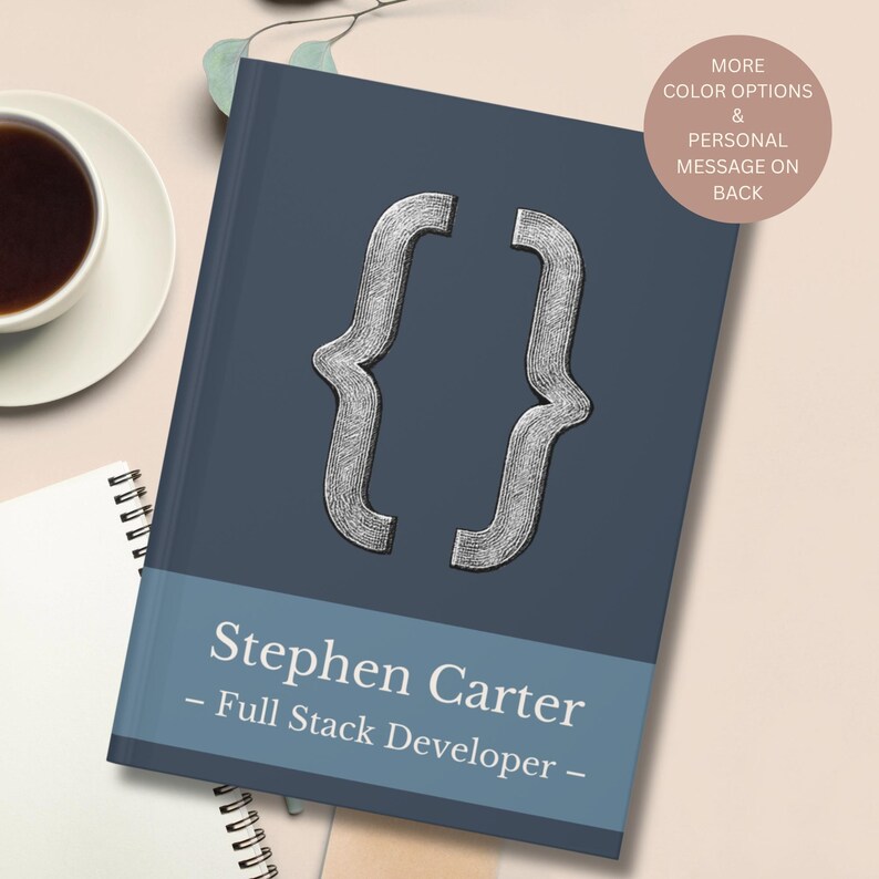 May include: A navy blue notebook featuring a silver bracket symbol. The notebook has a blue band with the name "Stephen Carter" and the title "Full Stack Developer". A coffee cup and a spiral notebook are in the background.