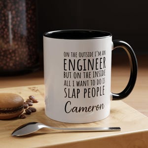 May include: White ceramic mug with a black interior and handle. The mug has the text "ON THE OUTSIDE I'M AN ENGINEER BUT ON THE INSIDE ALL I WANT TO DO IS SLAP PEOPLE Cameron" in black lettering.