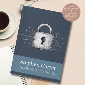 May include: A dark blue notebook with a silver padlock design and the text "Stephen Carter - CYBERSECURITY ANALYST -". The notebook has a white spiral binding and a coffee cup is visible in the background.
