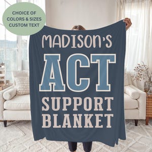 May include: A navy blue blanket with the text "MADISON'S ACT SUPPORT BLANKET" in white and light blue. The blanket is being held up, with a green circle in the upper left corner that says "CHOICE OF COLORS & SIZES CUSTOM TEXT".