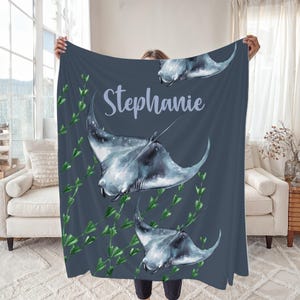 May include: A dark blue blanket featuring watercolor illustrations of manta rays and green seaweed. The name "Stephanie" is written in a light blue script across the blanket. The blanket is held up in front of a window and a couch.