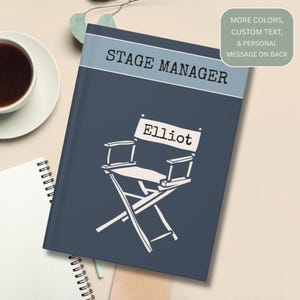 May include: A navy blue notebook with the text "STAGE MANAGER" and the name "Elliot" above a director's chair illustration. The notebook has a light blue band at the top and a green box with text on the back.