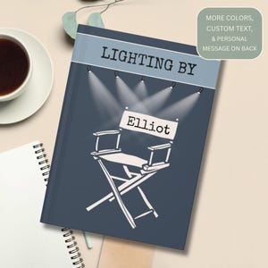 May include: A navy blue notebook with the text "LIGHTING BY" and a director's chair graphic with the name "Elliot". The notebook has a light blue band across the top and a white spiral notebook and a cup of coffee are in the background.