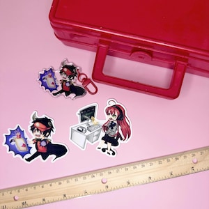 May include: Three anime-style acrylic charms and stickers featuring characters with red and black clothing. One charm has a red lobster clasp. A wooden ruler shows measurements in inches, and a large red plastic storage case is visible in the background.