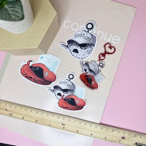 Könnte beinhalten: Eine Sammlung von drei Aufklebern und einem Acryl-Schlüsselanhänger mit Zeichentrickfiguren. Die Designs zeigen eine weiße Figur mit einer Kappe mit der Aufschrift 1148 und eine rote Figur. Ein Holzlineal zeigt die Skala in Zentimetern am unteren Rand.