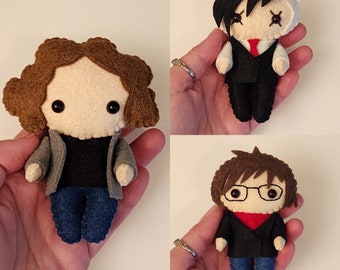 May include: Three felt plushies of cartoon characters. The first plush is brown with a black shirt and blue jeans. The second plush is black with a white shirt and black pants. The third plush is brown with a black shirt, red scarf, and blue jeans.