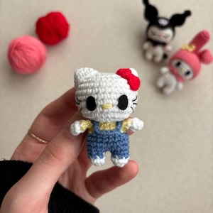 Può includere: Una bambola Hello Kitty di uncinetto bianca con una salopette di jeans blu e un fiocco rosso. La bambola è tenuta in una mano.