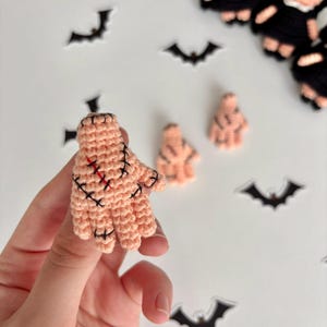 Puede incluir: Primer plano de una mano de ganchillo color melocotón con puntadas negras y detalles rojos. Varias figuras pequeñas de color melocotón y recortes con forma de murciélago se encuentran en el fondo. Las figuras parecen ser muñecas.