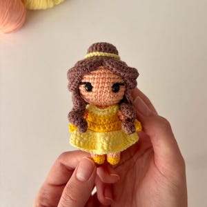 Könnte beinhalten: Handgefertigte Häkelpuppe in einem gelben Kleid mit braunen Haaren und einem gelben Stirnband. Die Puppe hat schwarze Augen und wird in den Händen einer Person gehalten. Wolle in Gelb- und Pfirsichtönen ist im Hintergrund.