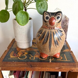 Puede incluir: Figura de búho de cerámica pintada a mano con cuerpo marrón, detalles negros y naranjas y pies rojos. El búho está posado sobre una caja de madera con libros debajo y una planta en maceta a la izquierda.