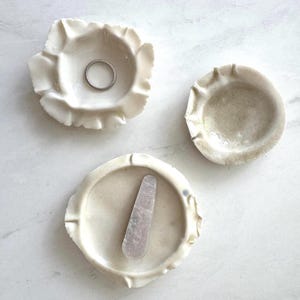 Puede incluir: Tres platos de cerámica en color blanco roto con bordes ondulados en forma de pétalos. Un plato contiene un anillo de plata fino, otro sostiene una pinza de pelo rectangular con superficie texturizada y el tercero está vacío y brillante.