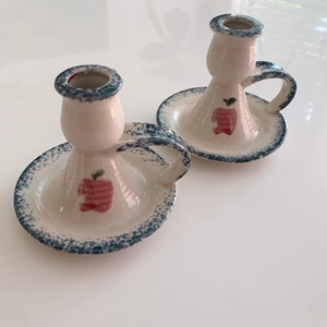 Puede incluir: Dos candelabros de cerámica blanca con motas azules y un diseño de manzana roja. Cada candelero tiene una pequeña asa.