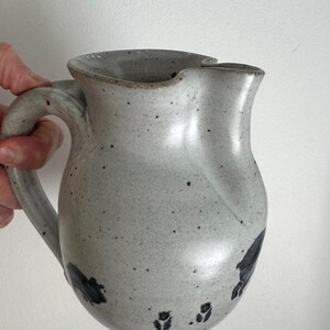 Puede incluir: Una jarra de cerámica de color gris claro con un acabado moteado y un asa curva. La jarra presenta un cuerpo ancho y redondeado, un borde acampanado y un pico vertedor distintivo. Pequeñas marcas pintadas oscuras y abstractas se ven en la base.