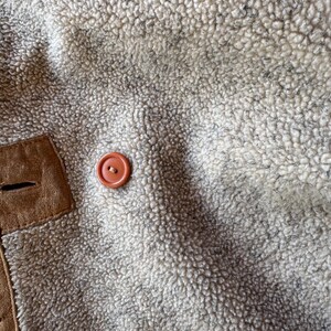 Pode incluir: Close-up do interior de um casaco de shearling bege e creme. Uma aplica&ccedil;&atilde;o de camur&ccedil;a castanha com uma casa de bot&atilde;o &eacute; vis&iacute;vel, juntamente com um bot&atilde;o redondo laranja. As bordas do casaco s&atilde;o aparadas com costuras decorativas.