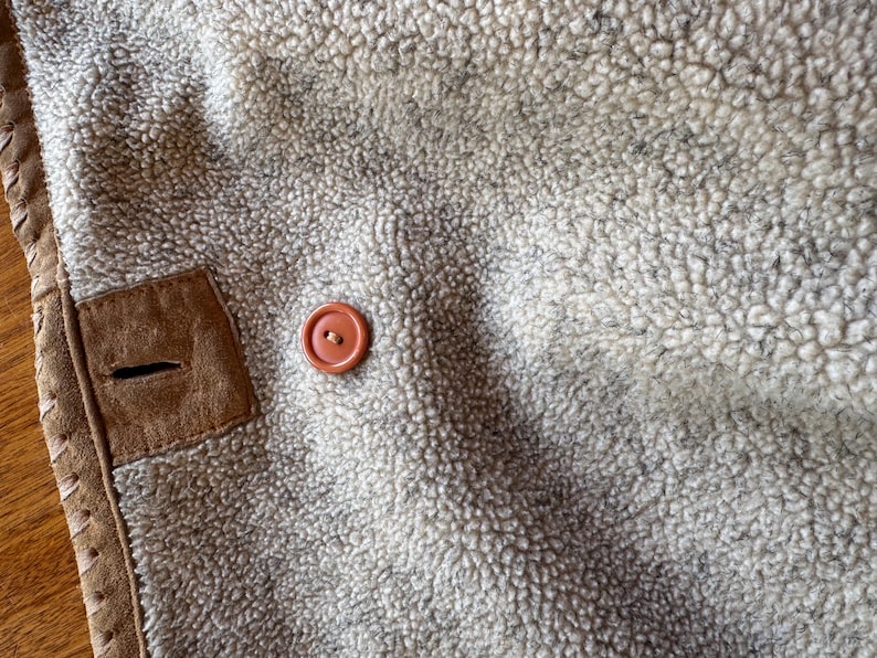 Pode incluir: Close-up do interior de um casaco de shearling bege e creme. Uma aplica&ccedil;&atilde;o de camur&ccedil;a castanha com uma casa de bot&atilde;o &eacute; vis&iacute;vel, juntamente com um bot&atilde;o redondo laranja. As bordas do casaco s&atilde;o aparadas com costuras decorativas.