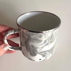 Puede incluir: Una taza de esmalte blanco con un diseño de mármol gris y un borde plateado. La taza tiene un asa.