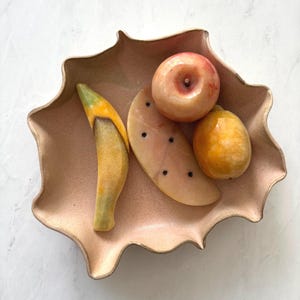 May include: A light pink, wavy-edged ceramic bowl containing four polished stone fruit carvings: a yellow and green banana, a pale pink watermelon slice with black dots, a round peach-colored apple, and a smooth, golden-yellow lemon.