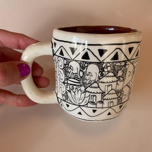 Pode incluir: Caneca de cerâmica branca com um design em preto e branco que apresenta padrões geométricos e figuras estilizadas. A caneca tem uma borda marrom e uma alça.