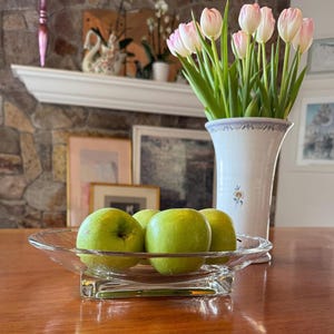 Puede incluir: Un cuenco de vidrio transparente con manzanas verdes sobre una mesa de madera. Un jarrón blanco con tulipanes de punta rosa y una repisa decorativa en el fondo.