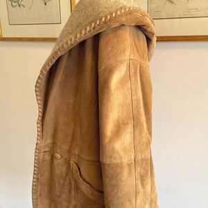 Pode incluir: Um casaco de camur&ccedil;a castanho com capuz e forro claro e texturizado. O casaco apresenta uma borda costurada ao longo das bordas, um bolso grande e um fecho de bot&atilde;o. O casaco &eacute; exibido em um manequim.