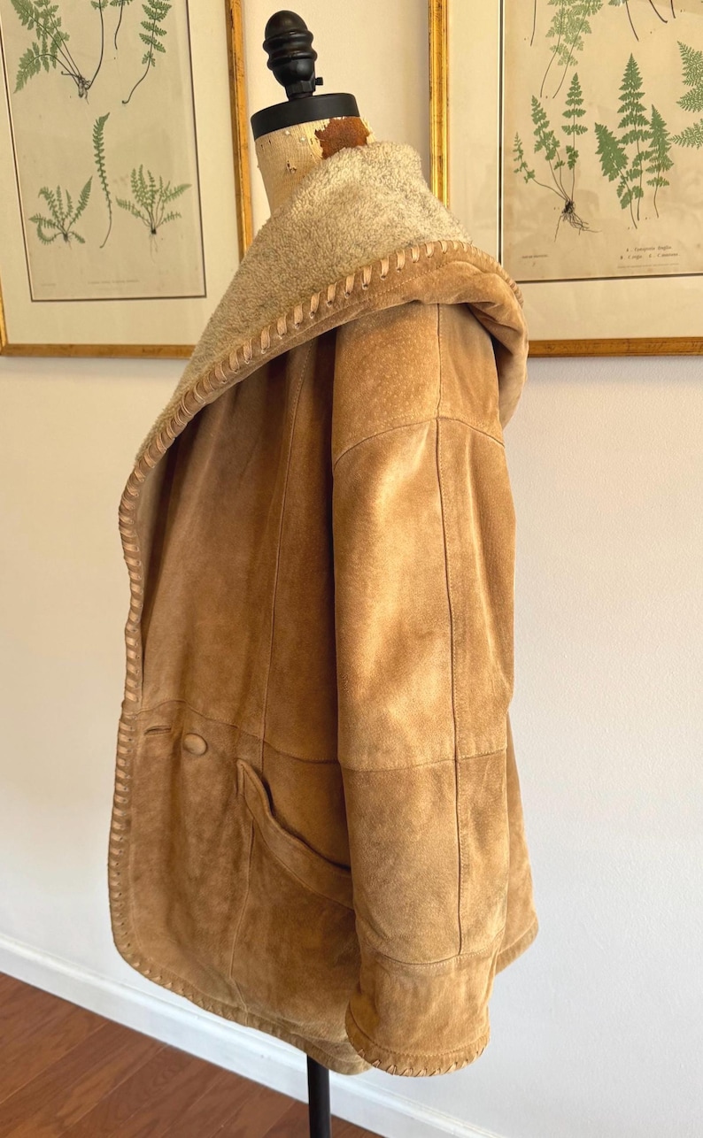 Pode incluir: Um casaco de camur&ccedil;a castanho com capuz e forro claro e texturizado. O casaco apresenta uma borda costurada ao longo das bordas, um bolso grande e um fecho de bot&atilde;o. O casaco &eacute; exibido em um manequim.