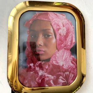 May include: A framed portrait featuring a woman with a pink floral head covering and matching lip color. The portrait is encased in a gold-colored, rectangular frame with rounded corners. The image has a soft, artistic quality.